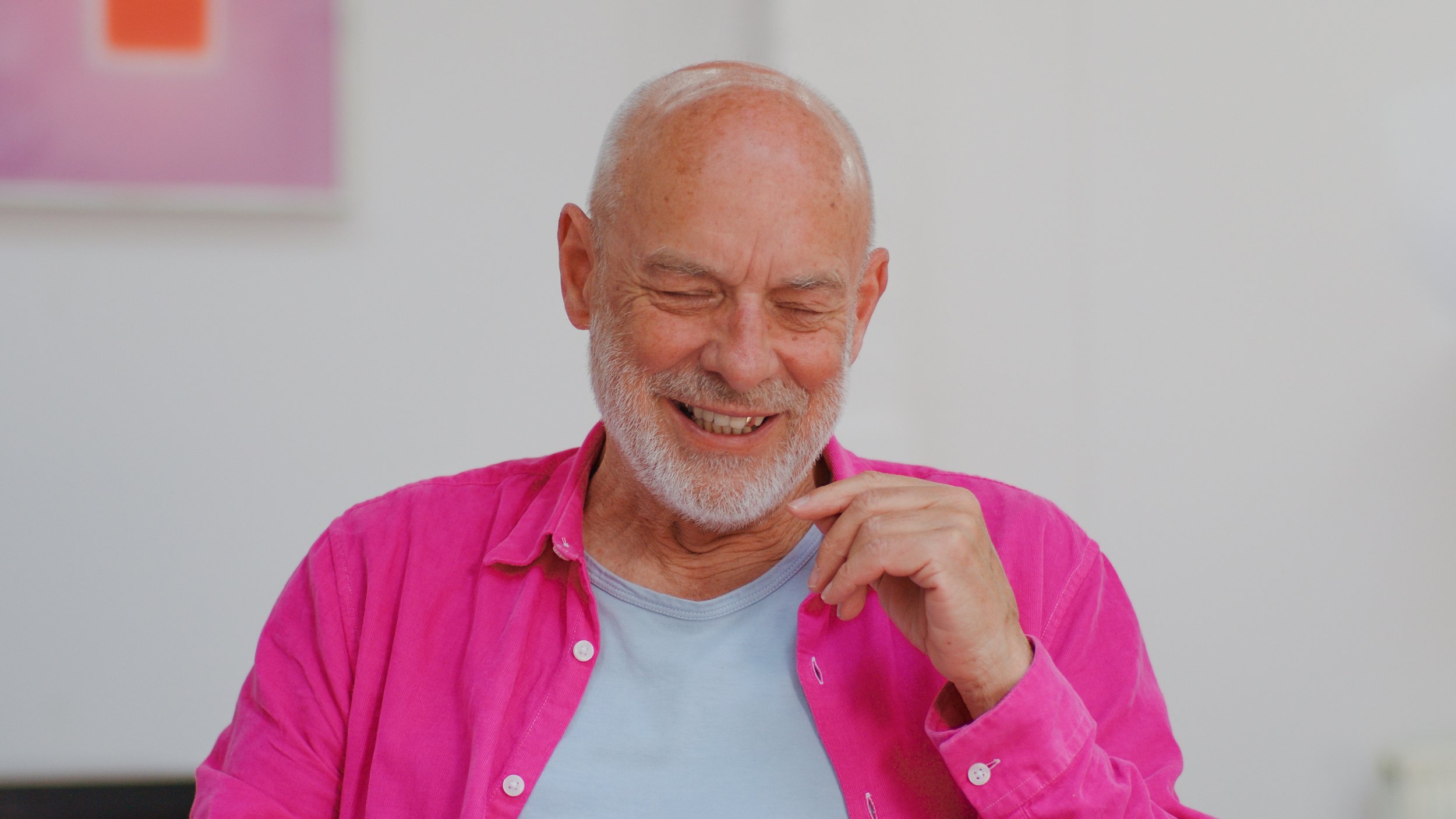 Brian Eno, wearing a pink jacket and a blue t-shirt, sits smiling with his eyes closed