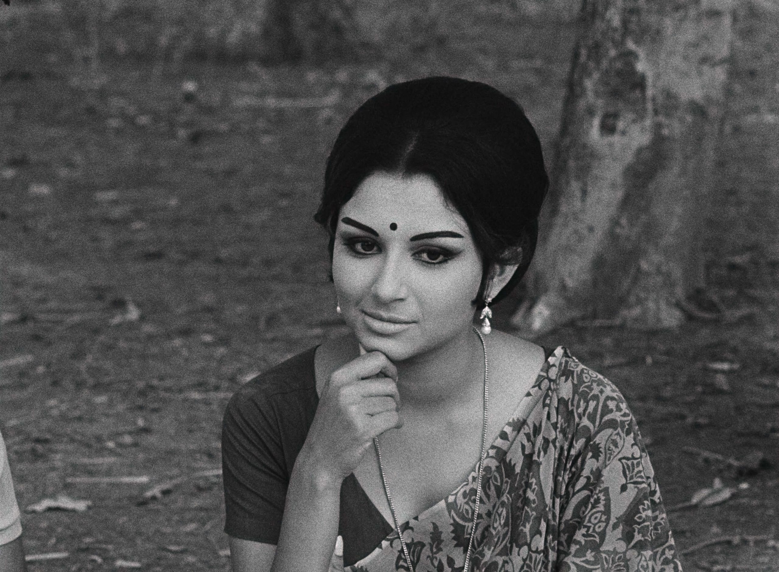 A woman with dark hair and a sari sits near a tree with her hand on her chin.
