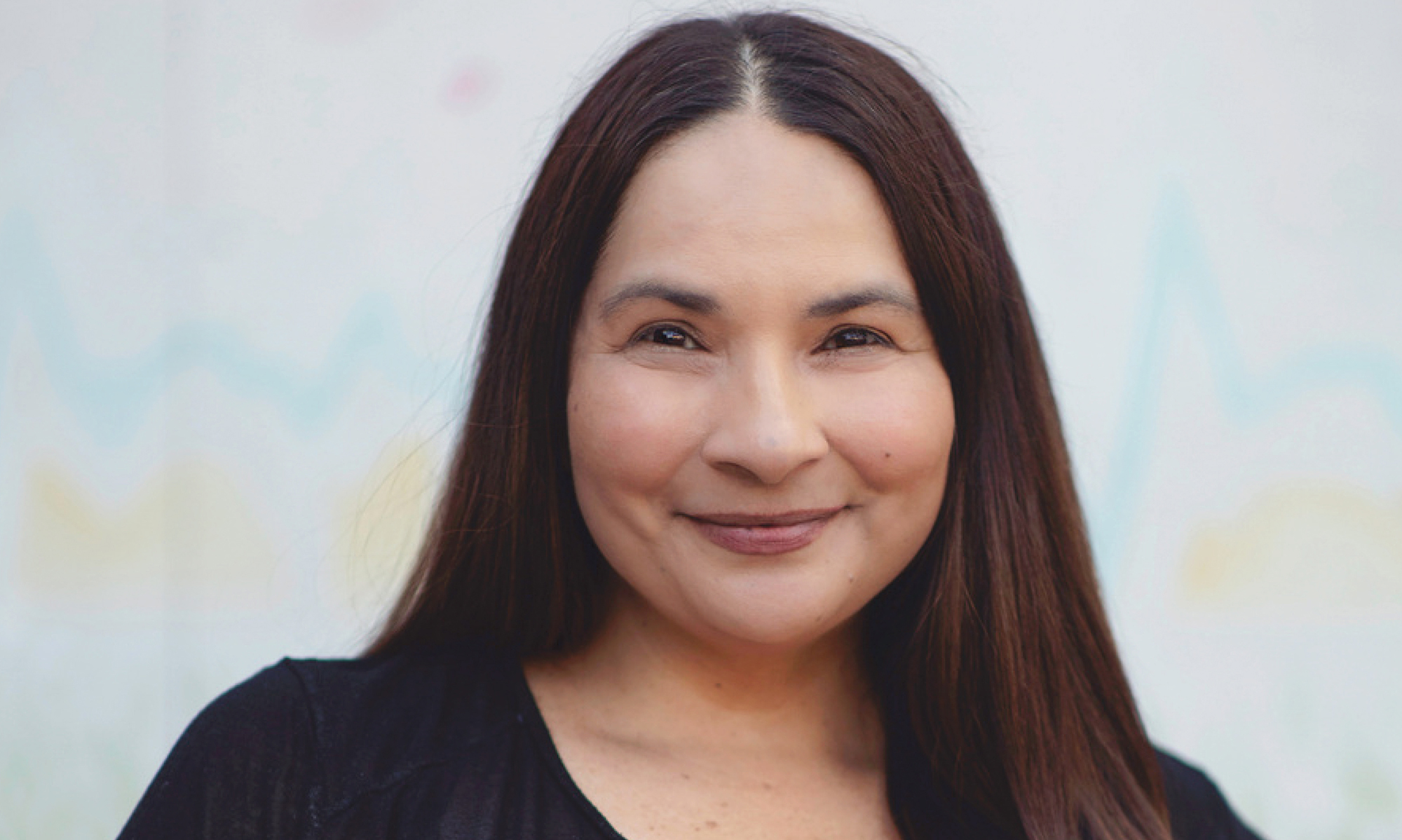 Headshot of Eva Thomas, writer and director of Nika and Madison. Brown Hair. Brown Eyes. Closed-mouth smile. Black, v-neck shirt. Blurred, white-ish background.
