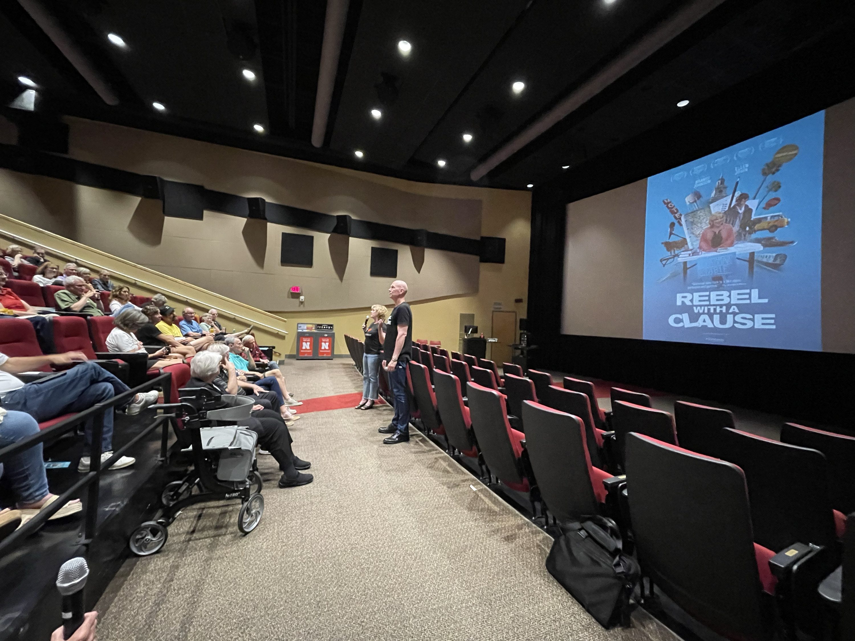 Photo of an auditorium, two people talking to people seated in the red chairs