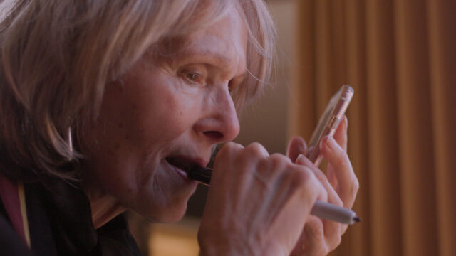 Older blonde woman looks at her phone as she holds it close to her face with a pen in her mouth.