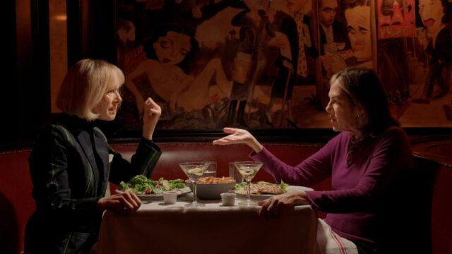 Two women engaged in an animated discussion over a meal in a restaurant booth. Wall in background is an illustrated mural.