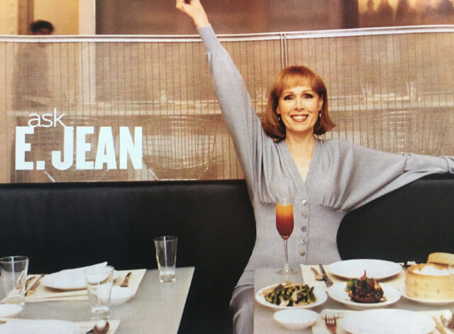 A happy woman smiles with outstretched arms while seated at a restaurant table holding plates of food.