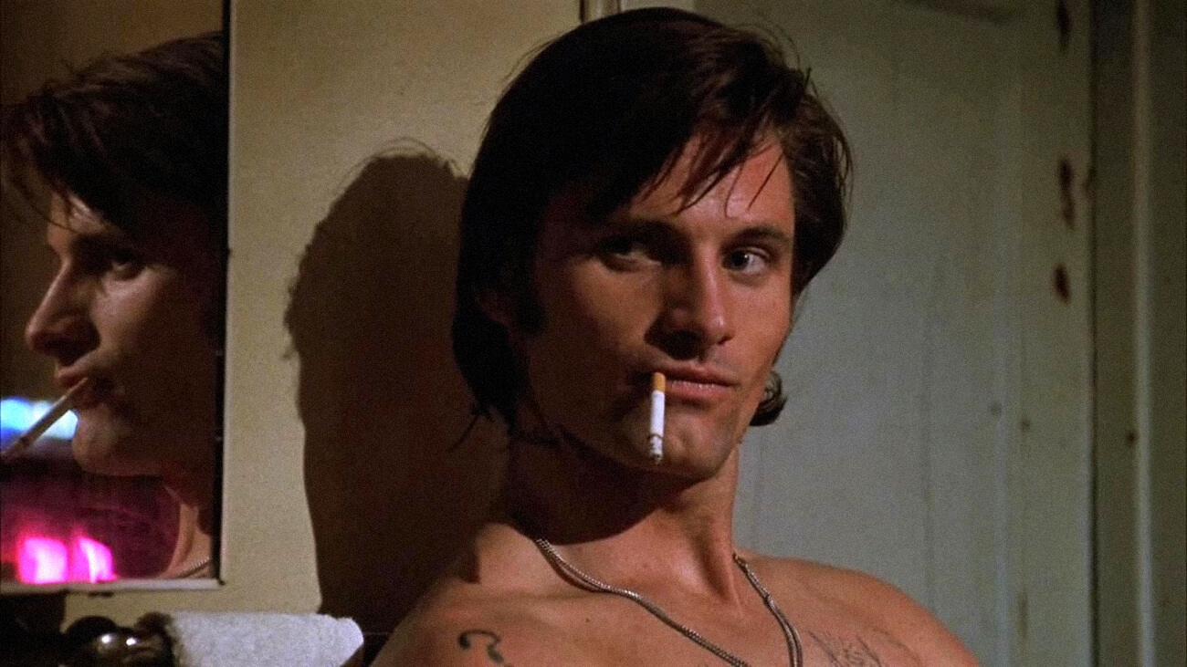 Shirtless white man with medium length hair holds cigarette in his moth with growing ash.