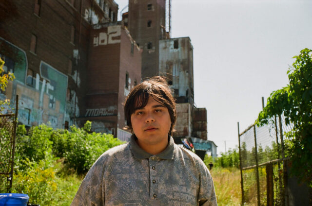 A scene from Mile End Kicks. Jesse (Isaiah Lehtinen) stands in front of a factory building.