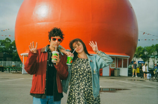A scene from Mile End Kicks. Chevy (Stanley Simons) and Grace (Barbie Ferreira) pose in front of an orange dome holding cups of orange juice.