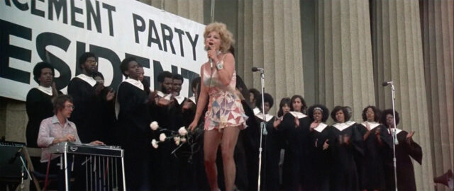 A scene from Nashville. A woman in a rhinestoned dress sings into a microphone with a gospel chorus behind her.