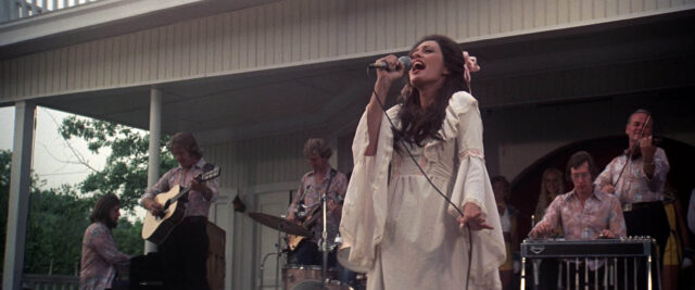 A scene from Nashville. A woman in a white dress sings into a microphone with a band playing instruments behind her.