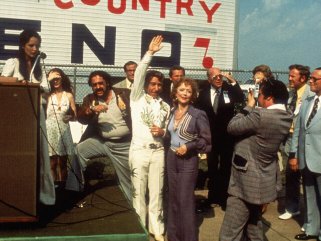 A scene from Nashville. A man in a white suit raises his hand to wave at a crowd.