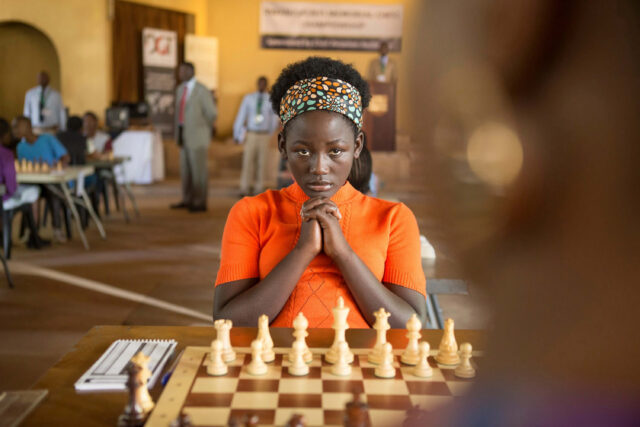A young woman clasps her hands and appears to be waiting intently in front of a chess board.