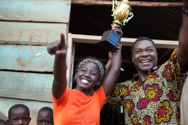 A young woman and a man celebrate with their hands in the air as the woman holds a trophy. Two children stand nearby.