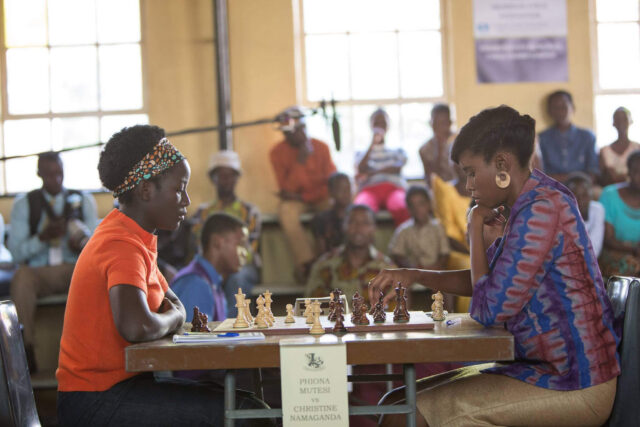 Two African women play chess in front of spectators.
