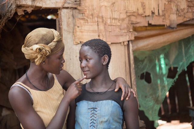 An African woman in a head wrap speaks to a younger woman in front of a dilapidated hut.