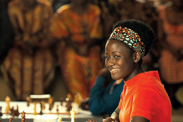 A smiling young African woman in brightly colored clothing sitting at a chess board.