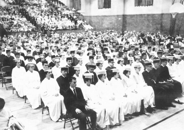 A gym of students in graduation caps and gowns, sitting in lines, and facing front.