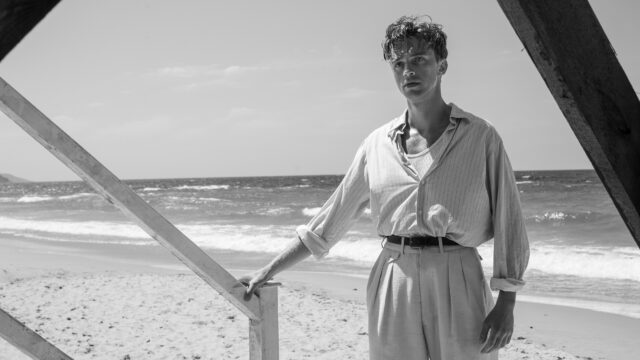 A man in 1940s clothing stands on a beach with the ocean behind him. Black and white.