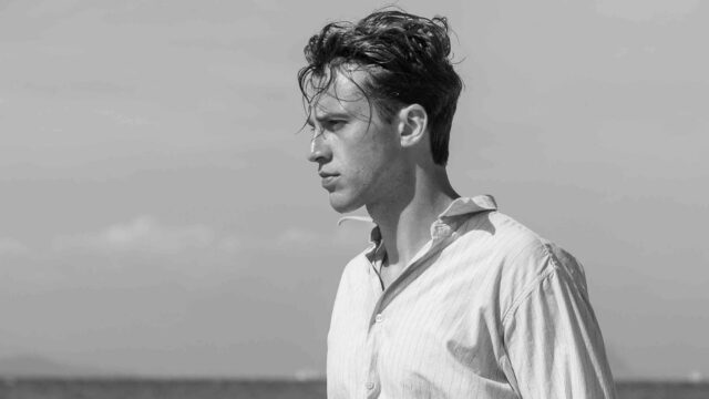 A black and white image of a man standing with tousled hair and a lose white shirt.