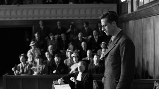 A man looks upset while standing in a court room while people in a jury box look on.