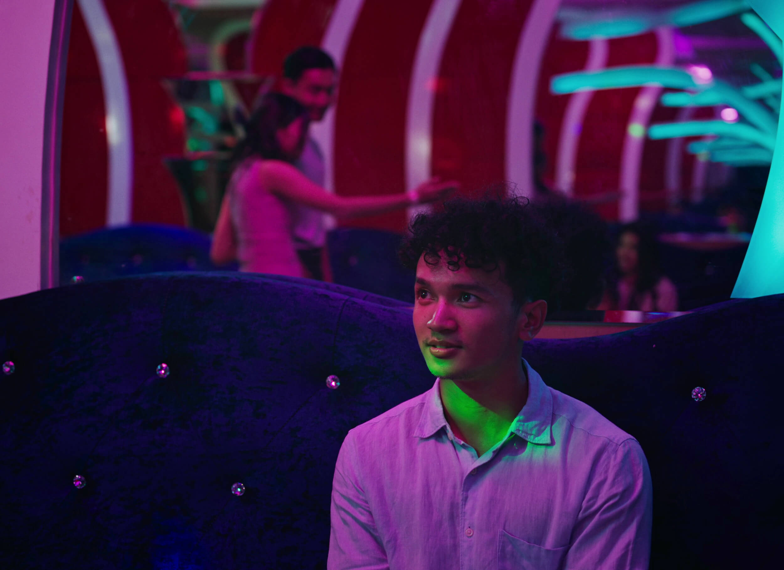 A scene from Grandma Nai Who Played Favorites. A young man with curly dark hair and a slight smile looks up from the blue velvet couch he's seated on with neon lights and people in the background.