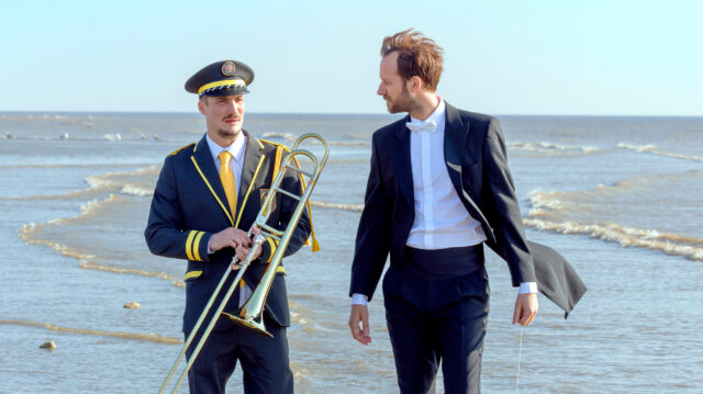 Pierre Lottin and Benjamin Lavernhe in a scene from Emmanuel Courcol’s THE MARCHING BAND. A man in a marching band uniform holding a trombone converses with a man in a tuxedo holding a conductor's baton as they stand on the shore of the sea.