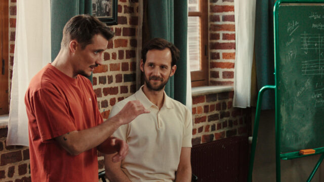 Pierre Lottin and Benjamin Lavernhe in a scene from Emmanuel Courcol’s THE MARCHING BAND. A man gestures as if conducting music while another man appears to admire him.