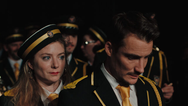 Sarah Suco and Pierre Lottin in a scene from Emmanuel Courcol’s THE MARCHING BAND. A woman in a marching band uniform peers at a man over his shoulder as he bows his head .