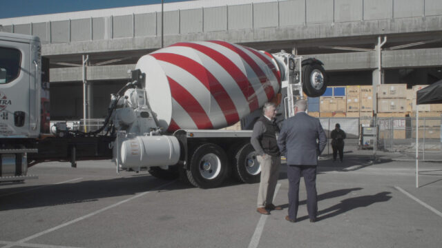 Two men stand in front of a cement truck in a scene from The History of Concrete.