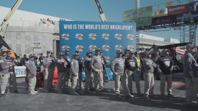 Bricklayers in grey shirts stand in line at a Best Bricklayer contest in a scene from The History of Concrete.