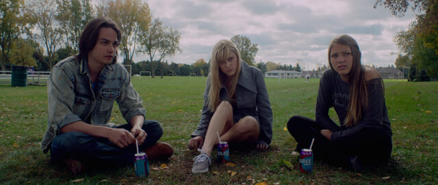 Three teens sit on the grass in a park in a scene from It Follows.