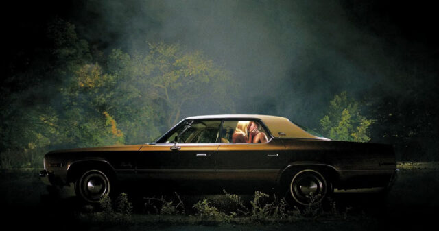 Two people kiss in the back seat of a car lit with dramatic lighting in a scene from It Follows.